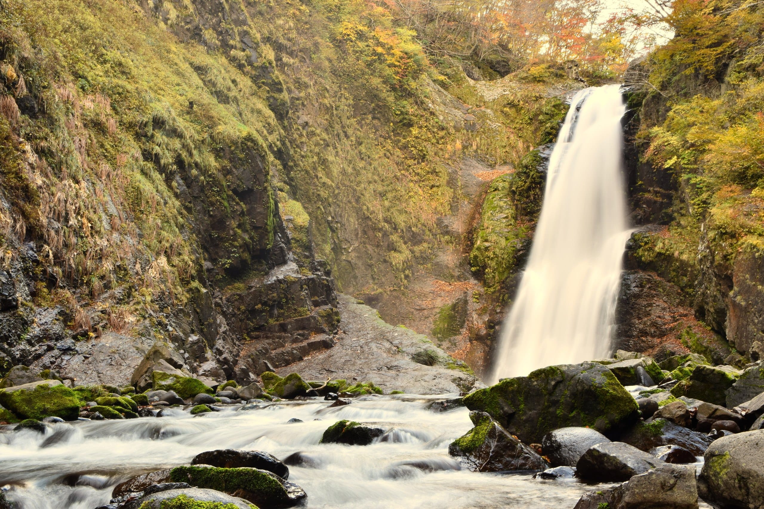 A tall waterfall pouring into a boulder-strewn stream in autumn