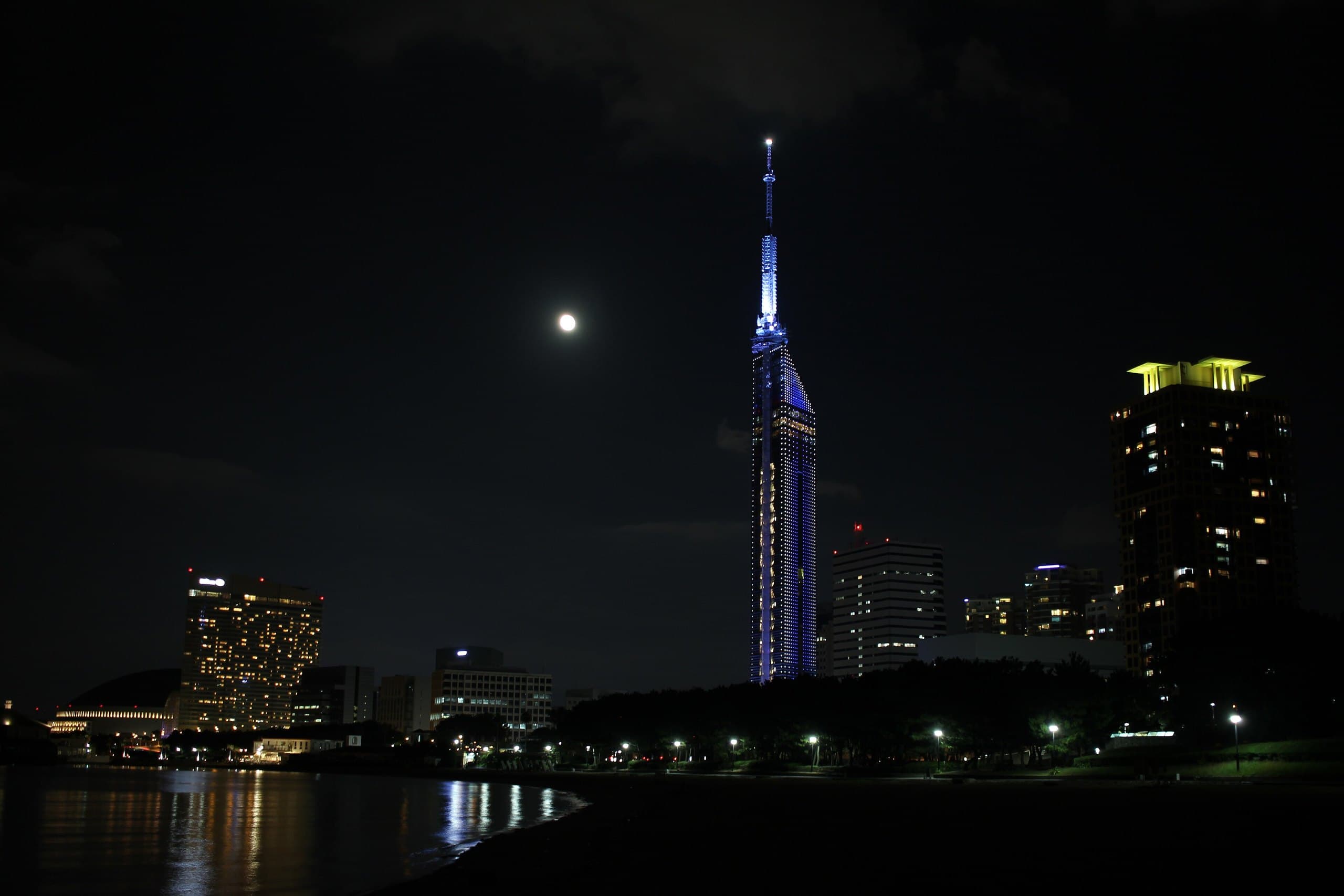 Fukuoka Tower lit blue at night with a full moon over the seaside district