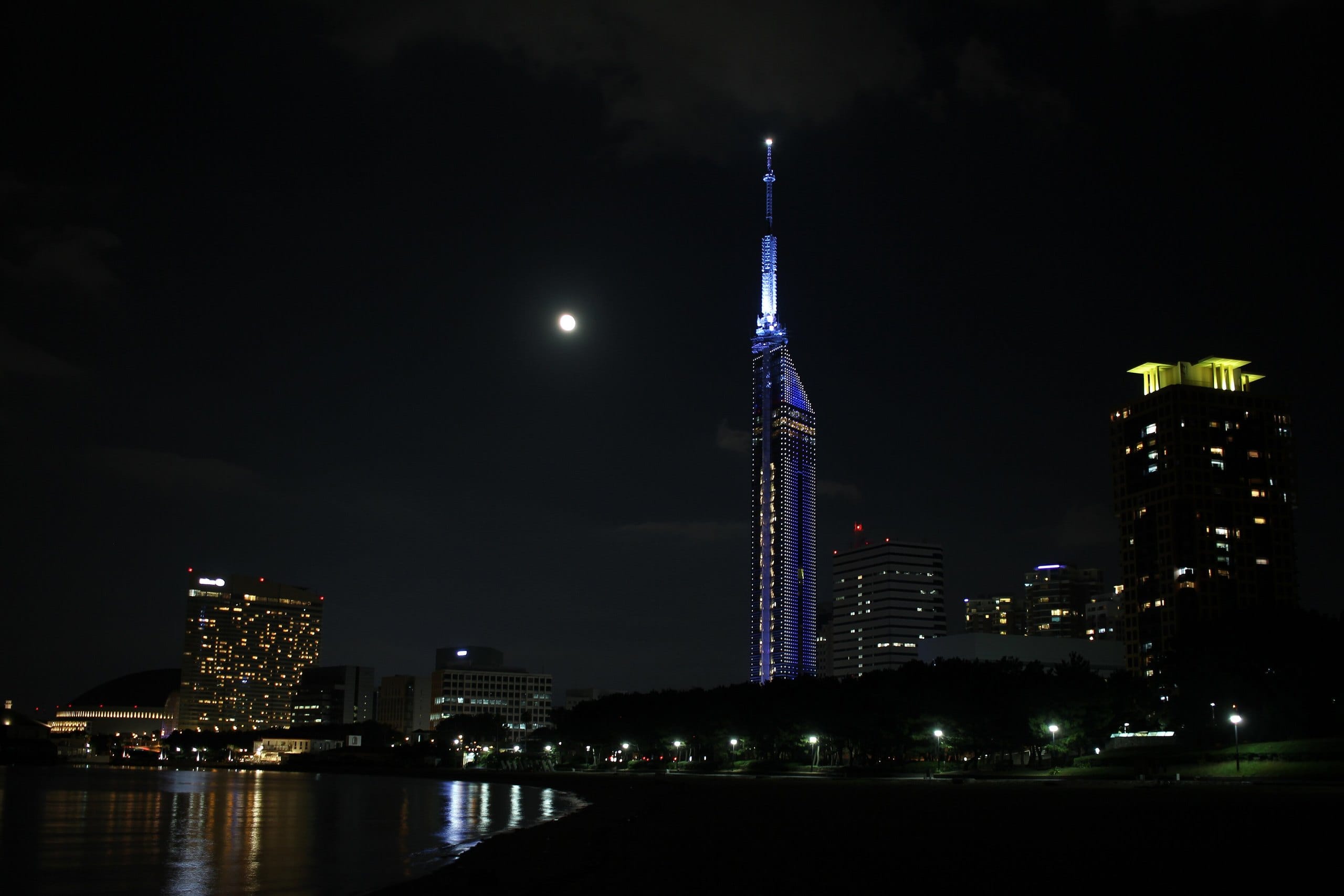 Fukuoka Tower lit blue at night with a full moon over the seaside district