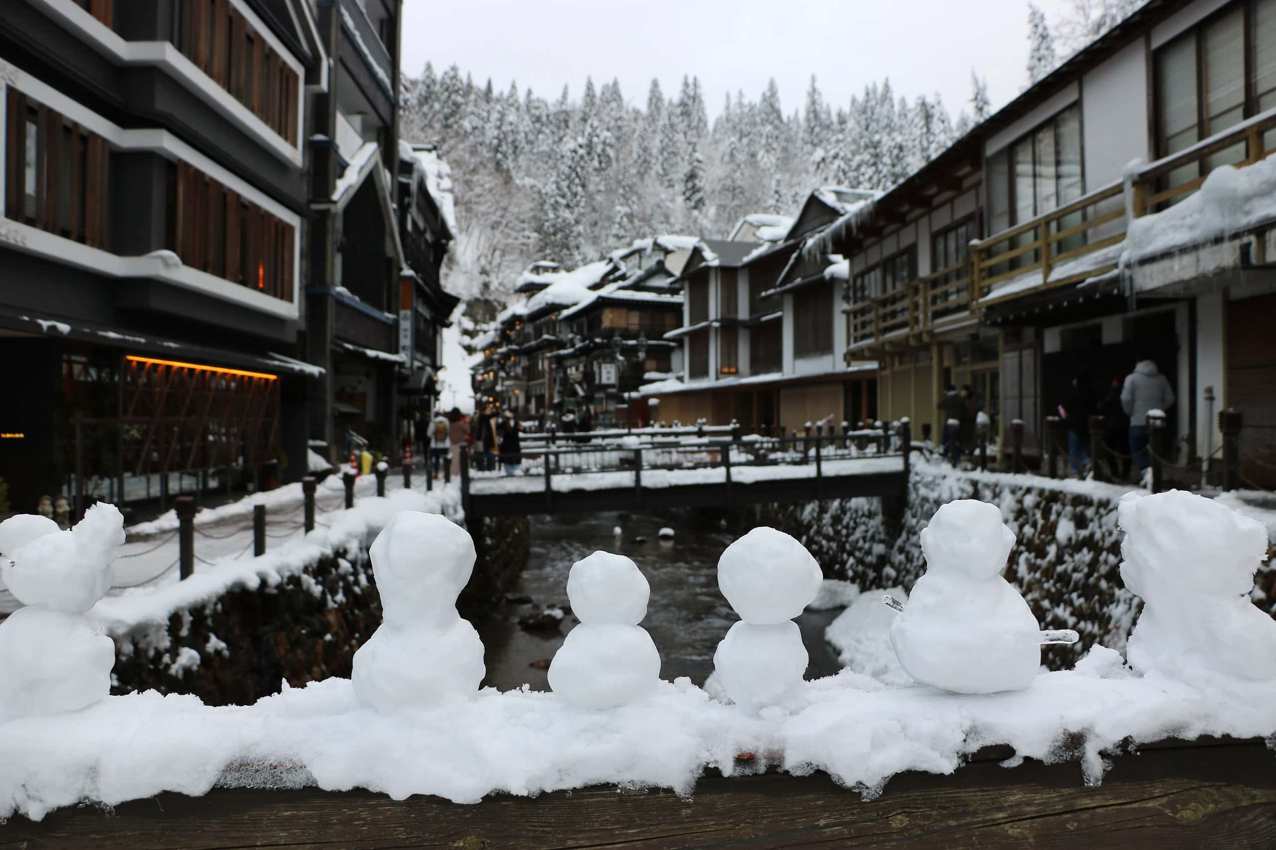 Row of small snowmen on a railing with snowy Ginzan Onsen buildings beyond