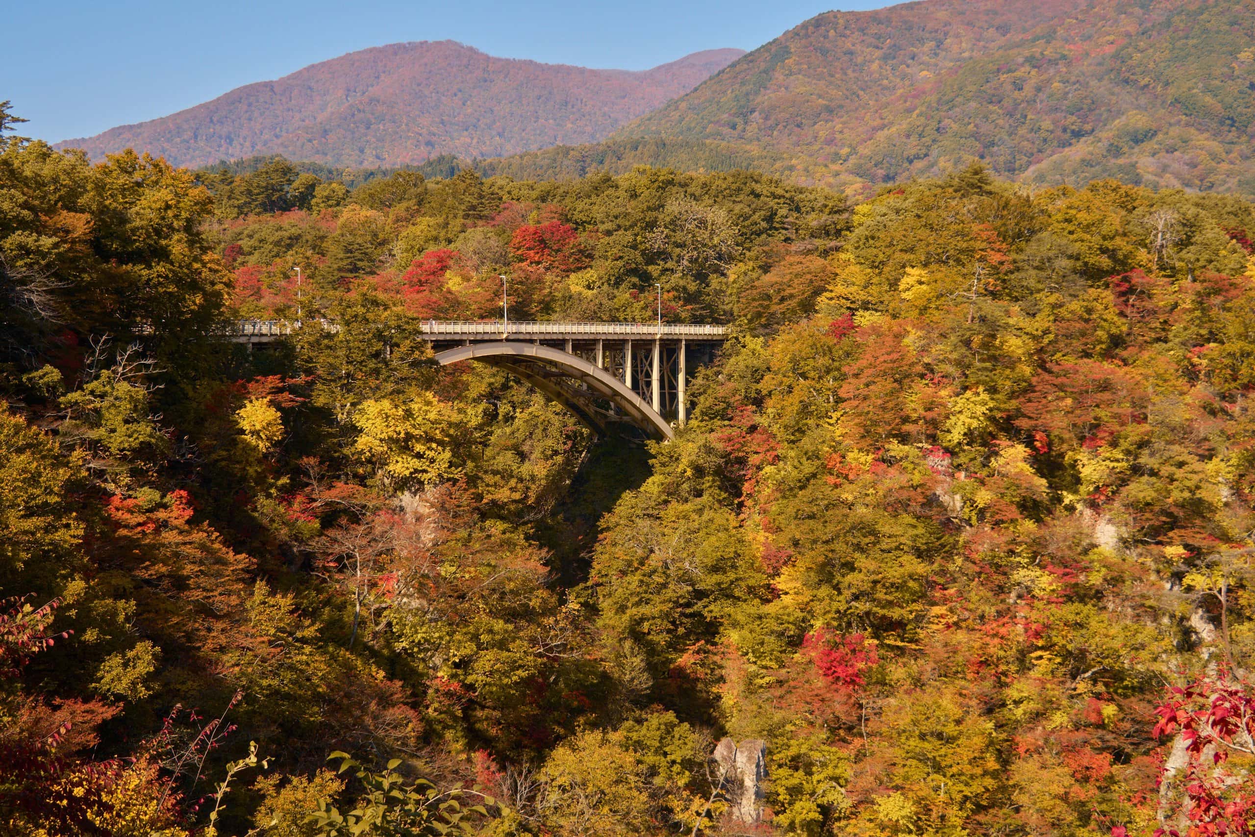Arched road bridge spanning Naruko Gorge amid red and gold autumn forest