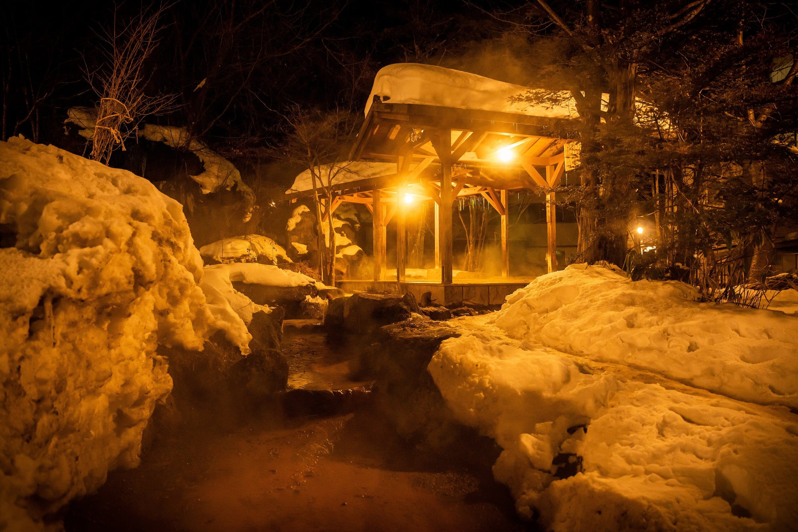 Snowbanks and a lantern-lit wooden bath shelter at night in Okuhida