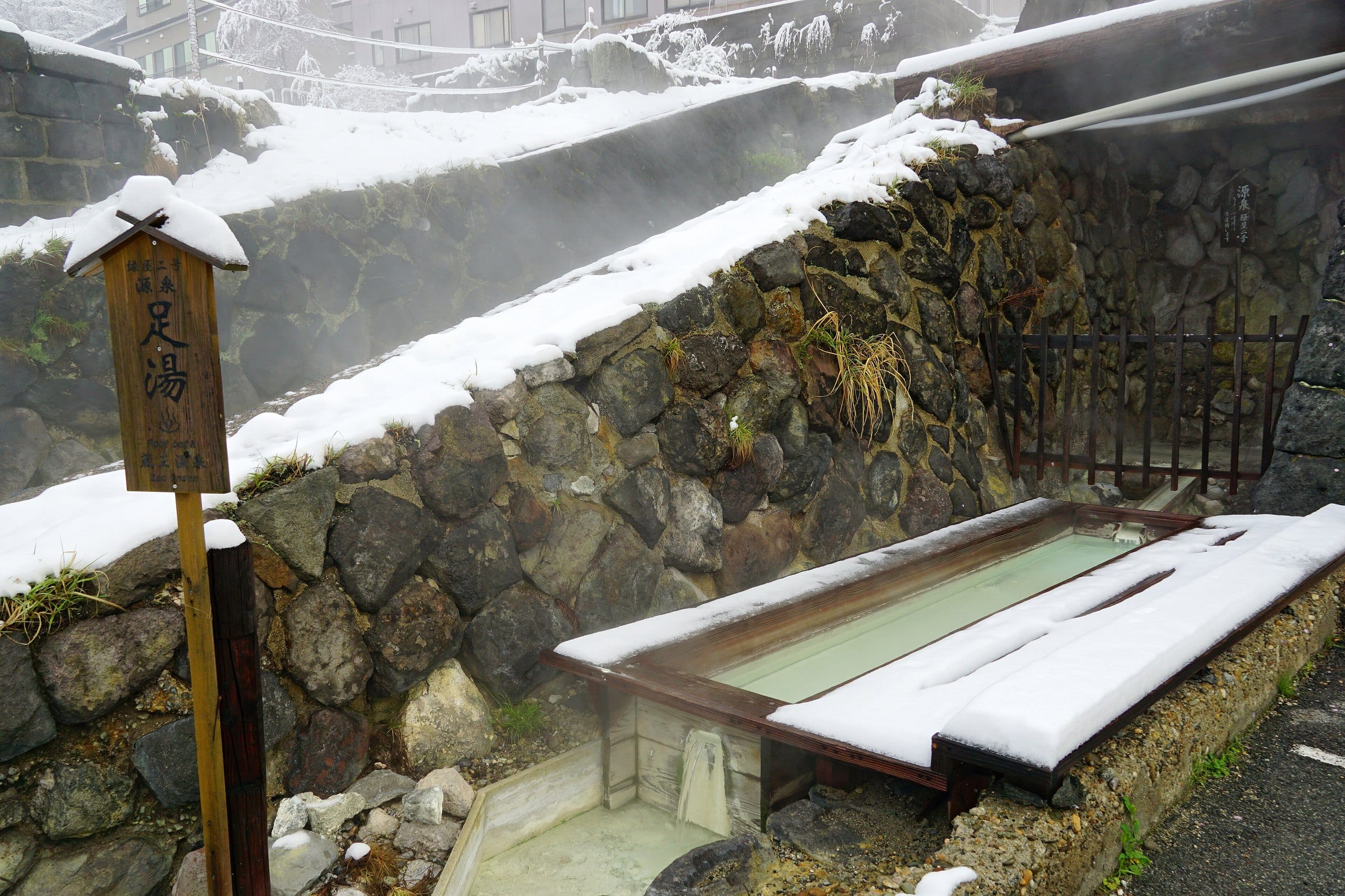 A snow-dusted public foot bath beside a stone wall with hot spring steam