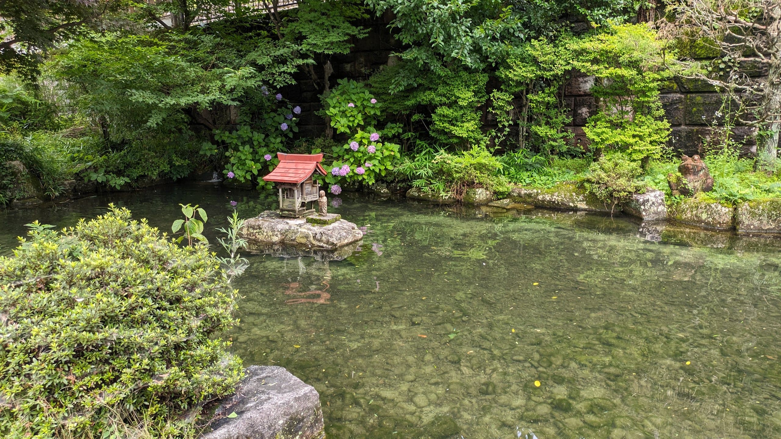 A small red-roofed shrine on a stone in a clear garden pond surrounded by greenery